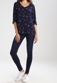 Blouse marine avec un col en V, des manches retroussées et des motifs blancs, rouges et bleus d'instruments de musique et d'étoiles. Assortie à un jean foncé et des baskets blanches.