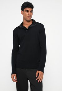 Schwarzer Strickpullover mit Viertelreißverschluss-Kragen, der aus strukturierter Stoff besteht und eine taillierte Passform hat. Akzente sind ein robuster Reißverschluss und seitliche Saumdetails.