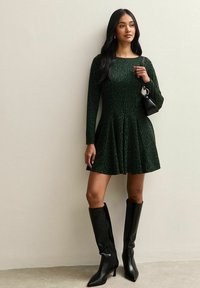 Donna che indossa un abito leopardato verde scuro con maniche lunghe, stivali neri con tacco fino al ginocchio e tiene in mano una piccola borsa nera.