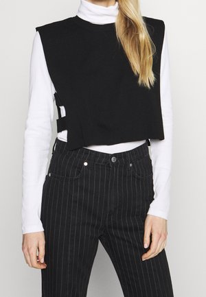 Frau trägt eine schwarze ärmellose, bauchfreie Weste über einem weißen Rollkragenpullover und schwarze Nadelstreifenjeans, dargestellt von der Taille aufwärts.