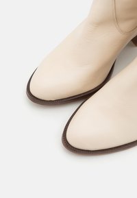 Botas de cuero beige hasta el tobillo con punteras puntiagudas y suelas texturizadas marrones. Presentan costuras sutiles a lo largo del costado para un detalle adicional.