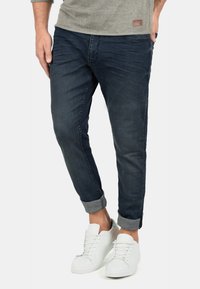 Tamne tamne traperice tamnoplave boje s preokrenutim manžetama, uparene s bijelim tenisicama. Glatka tekstura, slim fit dizajn i minimalistički detalji šivanja.
