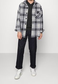 Chemise en flanelle à carreaux noirs et blancs portée sur un pull noir à col en V, associée à un pantalon bleu marine et des baskets blanches. Détail de poche notable sur la chemise.