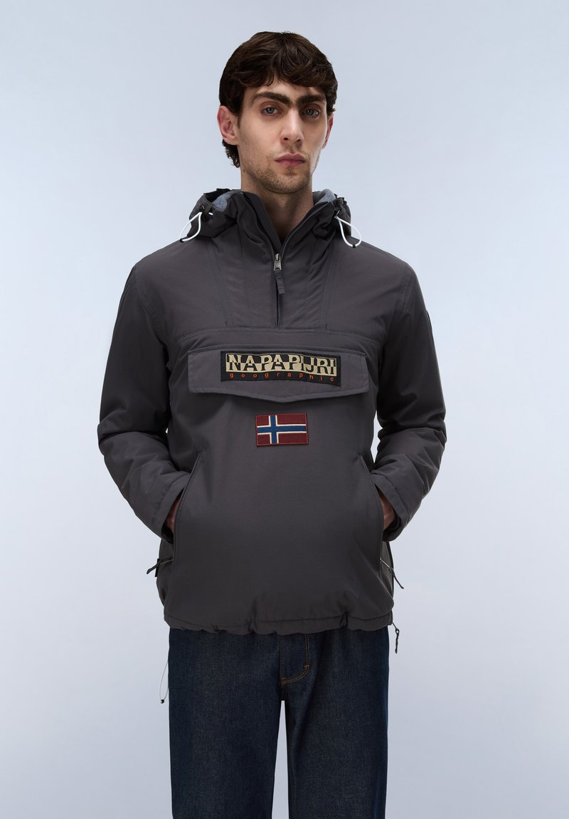 Junger Mann trägt eine dunkelgraue Napapijri Kapuzenjacke mit Fronttasche und norwegischem Flaggenaufnäher, steht vor einem schlichten hellen Hintergrund.