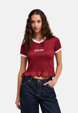 Femme aux longs cheveux ondulés portant un crop top rouge smocké avec le texte « "Ruca" » et un jean foncé taille haute, debout devant un fond uni.