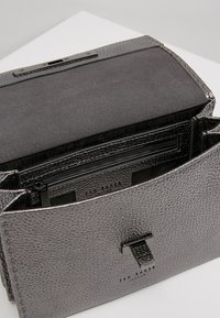 Szara torebka ze skóry o fakturze, z magnetycznym zapięciem na klapę. Wnętrze gładkie, ciemne, z kieszonką na zamek, oznaczone marką "Ted Baker".
