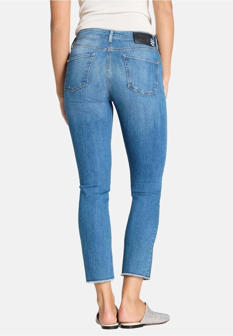 Femme portant un jean skinny court bleu clair avec des ourlets effilochés et des ballerines à enfiler à motifs, vue de dos sur un fond uni.