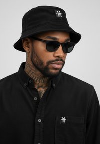 Black bucket hat with textured fabric, featuring a logo patch. Worn with black button-up shirt, showcasing a pocket and subtle pattern.