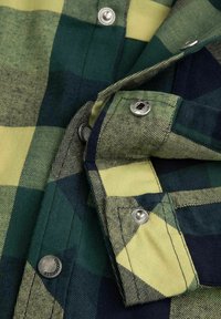 Tkanina w zieloną i żółtą kratę flanelową z widocznymi guzikami i zatrzaskami. Posiada teksturowaną powierzchnię oraz kontrastowy kołnierz.