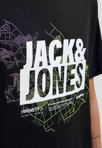 Sort t-shirt med stort hvidt tekst "JACK & JONES" og neon grønne og lilla bykort design som baggrundselementer.