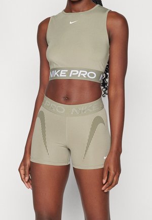 Femme portant un débardeur court Nike Pro vert olive et un short de sport taille haute assorti avec des détails de logo blanc, debout devant un fond uni.