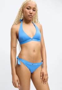 Νεαρή γυναίκα με μακριά ξανθά πλεξούδες, ντυμένη με μπλε μπικίνι Tommy Hilfiger με τοπ halter και σλιπ με δεσίματα στο πλάι, στέκεται μπροστά σε λευκό φόντο.