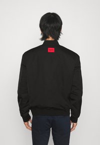 Giacca bomber nera con un patch rosso sul retro, polsini a costine e una texture liscia. Design casual con una vestibilità comoda.