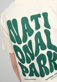 T-shirt en coton crème avec un grand texte vert en gras qui dit "PARC NATIONAL" dans une typographie stylisée et organique.