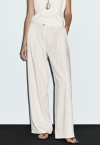 Pantalons large en beige clair, fabriqués dans un tissu doux, avec des poches avant et une fermeture à bouton. Associés à un haut simple et des sandales.