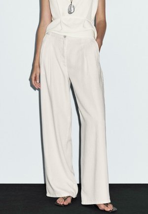 Weite, hellbeige Hosen aus weichem Stoff, mit Fronttaschen und Knopfverschluss. Kombiniert mit einem schlichten Oberteil und Sandalen.