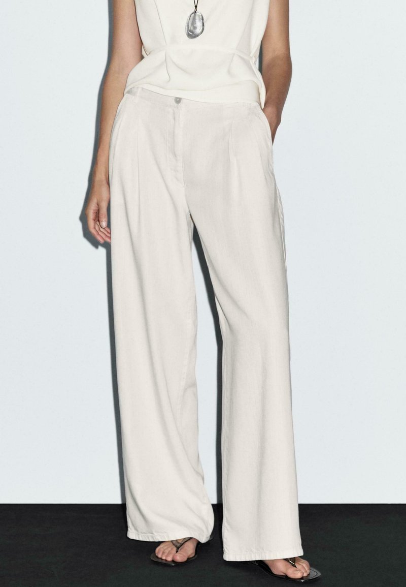 Pantalons large en beige clair, fabriqués dans un tissu doux, avec des poches avant et une fermeture à bouton. Associés à un haut simple et des sandales.
