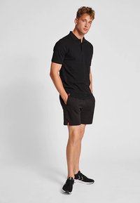 Sort poloshirt lavet af bomuld, kombineret med sorte shorts med røde accenter, båret med sorte sportssko. Modellen står i en neutral positur.