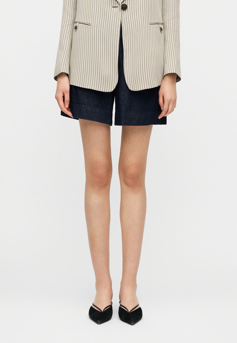 Γυναίκα που φοράει μαύρες μυτερές γόβες, σκούρο  μπλε σορτς και μπλέιζερ με μπεζ ρίγες, σε ασπρόμαυρο φόντο.