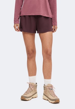 Person iført vinrøde shorts, en langærmet top, hvide sokker og beige vandrestøvler, der står mod en ensfarvet baggrund.