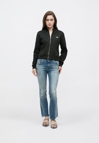 Sweatshirt preta com fecho de correr, design ajustado e punhos canelados, combinada com jeans azul claro flare. O modelo usa sandálias pretas sobre um fundo simples.