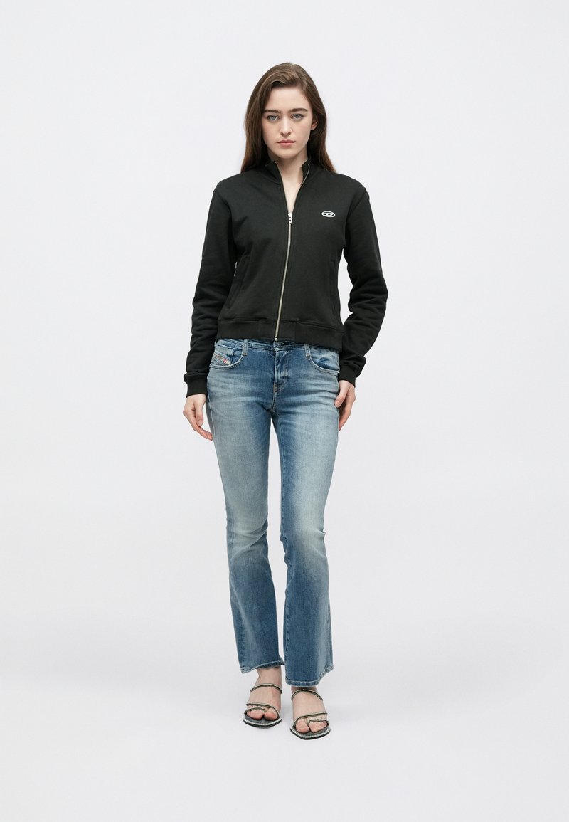 Sort lynlåshættetrøje med tætsiddende design og ribstrikkede ærmeafslutninger, kombineret med lyseblå vide jeans. Modellen har sorte sandaler på en ensfarvet baggrund.