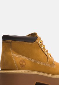 Tan suède enkelboot met een bruine leren kraag, gele veters, witte stiksels en een logo dat op de zijkant is geperst. Waterdicht ontwerp.