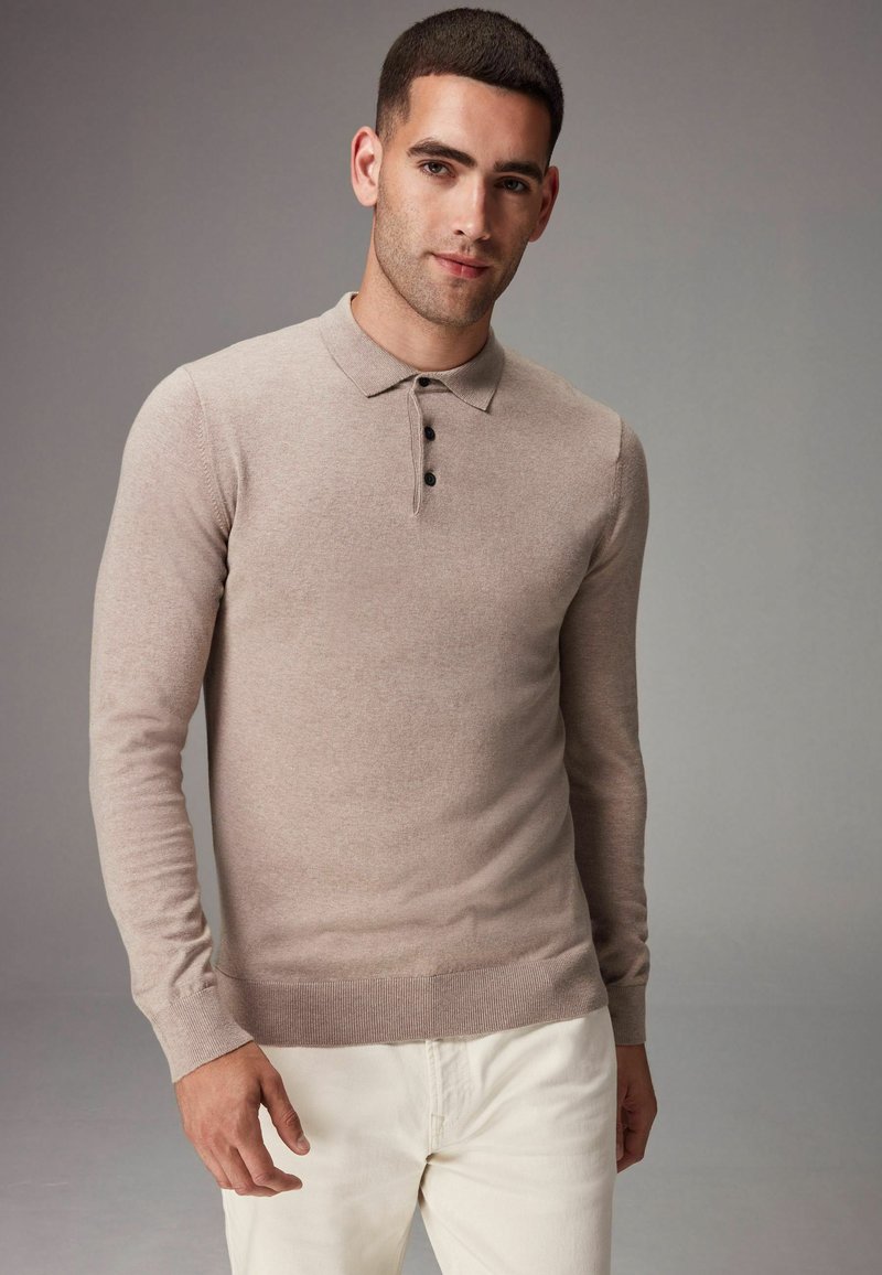 Polo shirt bej cu mânecă lungă, realizat din material tricotat, având un guler plat și un sistem de închidere cu trei nasturi. Purtați cu pantaloni de culoare deschisă.