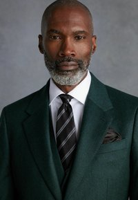 Traje de lana en verde oscuro con una camisa blanca y una corbata negra a rayas. Pañuelo de bolsillo negro con un patrón sutil. Barba gris bien cuidada.