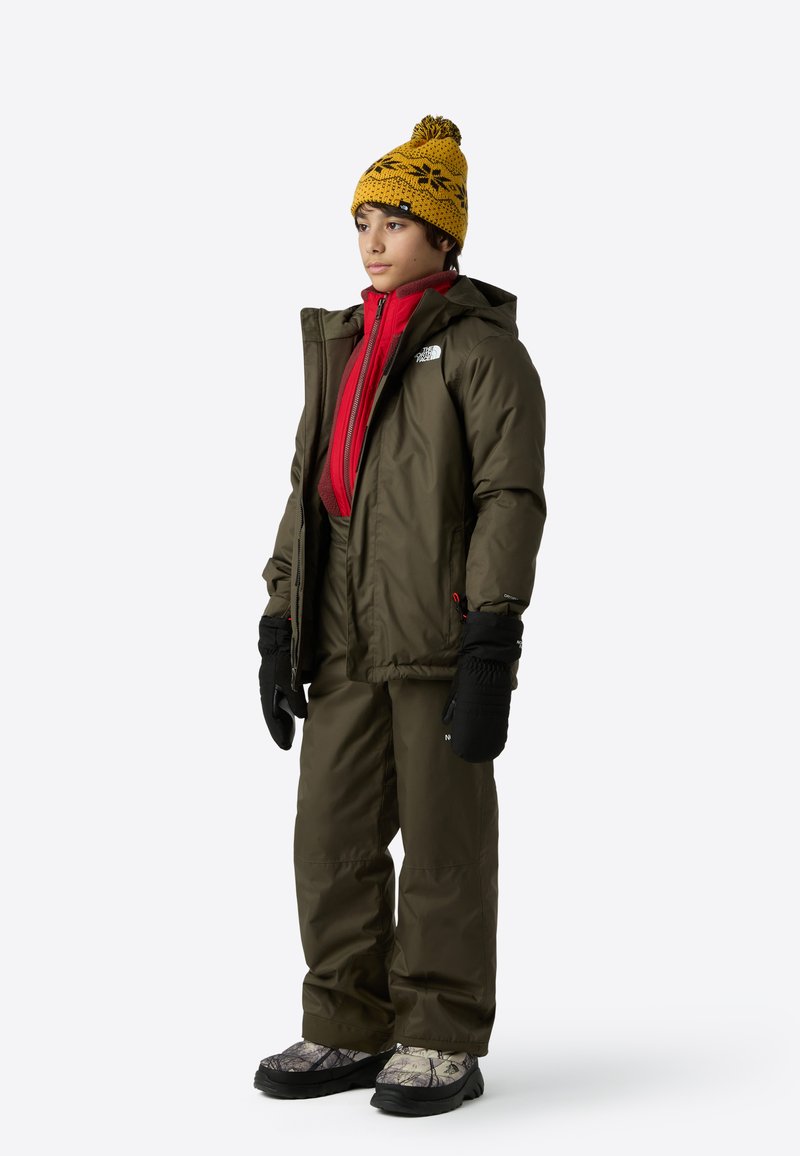 Insulated winter outfit featuring an olive green jacket and pants, red inner layer, black gloves, and a yellow patterned beanie. Textured boots.