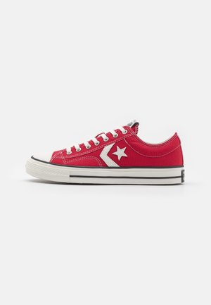 Roter niedriger Converse-Sneaker mit weißem Sternlogo, weißen Schnürsenkeln und weißer Gummisohle mit schwarzen Streifen auf weißem Hintergrund.