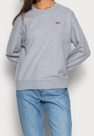 Vrouw draagt een lichtgrijze Levi's trui met een klein rood logo op de borst en lichtblauwe spijkerbroek, staand met haar handen achter haar rug.
