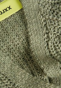 Gebreid groen trui stof met een gestructureerd patroon; bevat een glad stuk, toont dikke garens en een zichtbaar merklabel in geel.