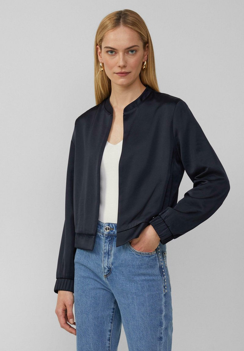 Veste bomber en satin bleu marine avec un col rond, des poignets côtelés et une coupe courte. Portée sur un haut blanc et associée à un jean bleu clair.
