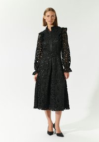 Schwarzes Spitzenkleid mit aufwendigen floralen Ausschnitten, tailliertem Oberteil, langen Ärmeln mit Rüschendetails und knielangem Rock.