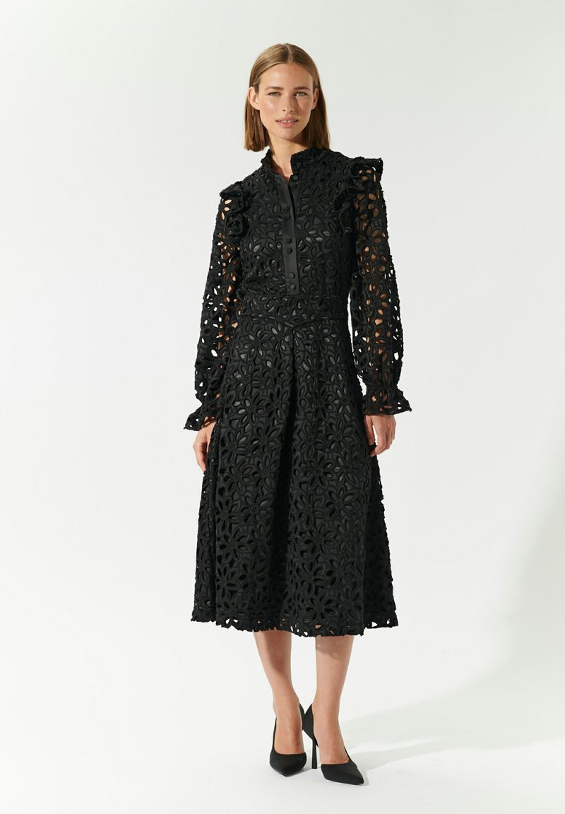 Vestido negro de encaje con intrincados recortes florales, corsé ajustado, mangas largas con detalles de volantes y falda a la altura de la rodilla.