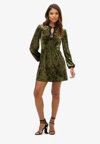 Femme portant une robe mini à manches longues en velours vert olive avec un décolleté en trou de serrure et des sandales à talons hauts noires à lacets sur fond blanc.