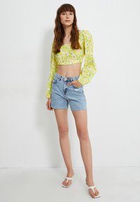 Żółta kwiatowa bluzka crop z długimi bufkami, w zestawie z szortami z wysokim stanem w stylu distressed. Modelka nosi białe sandały.