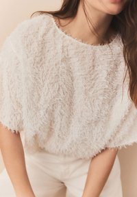 Top bianco peloso caratterizzato da un design loose e ampio con collo rotondo e maniche corte, realizzato in materiale testurizzato. Abbinato a pantaloni bianchi.