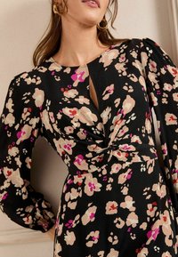 Rochie florală din material negru, cu modele în nuanțe de crem, roz și mov. Detaliu cu nod la talie și mâneci bufante, cu un decolteu în formă de V ușor.