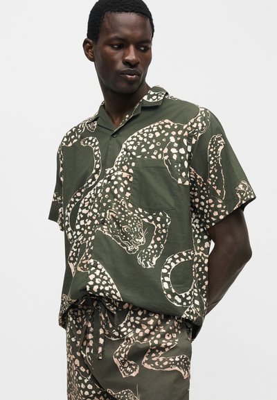 Άνδρας που φοράει πράσινο πουκάμισο με κοντό μανίκι και παντελόνι με μπεζ λεοπάρ σχέδιο, στέκεται με τα χέρια πίσω από την πλάτη κοιτώντας προς τα κάτω.