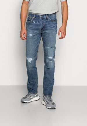 Homme portant un jean bleu délavé et des baskets grises debout devant un fond blanc uni.