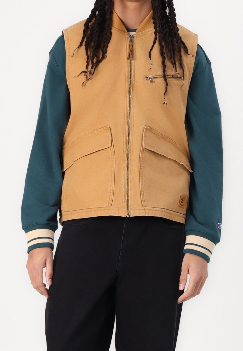 Personne portant un gilet utilitaire couleur fauve avec fermeture éclair et poches à l'avant, par-dessus un sweat-shirt à manches longues turquoise et un pantalon noir, debout devant un fond blanc.