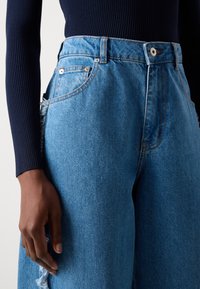 Hellblaue Denim-Jeans mit hoher Taille, versehen mit einem used Look und klassischem Fünf-Taschen-Design, kombiniert mit einem dunkel gerippten Oberteil.