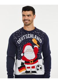 Brede gebreide trui met een donker marineblauwe basis, versierd met een feestelijke kerstman die een biertje en een voetballen vast heeft, met de tekst "DEUTSCHLAND" erboven.
