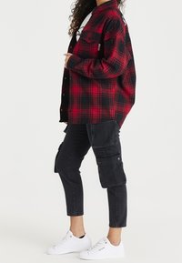 Chemise en flanelle à carreaux rouges et noirs, design à boutons, portée par-dessus un t-shirt graphique blanc. Associée à un pantalon cargo noir et des baskets blanches.