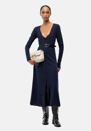 Donna che indossa un lungo vestito blu navy con bottoni e cintura nera, stivali neri, che tiene in mano una borsa crema con accessori dorati su sfondo bianco.