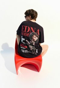 T-shirt noir oversize avec un graphisme d'un personnage portant des lunettes. Le texte inclut "EDNA MODE" et des règles de mode. Porté avec une chaise rouge.