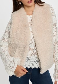 Gilet beige soffice con una texture morbida, caratterizzato da un'apertura frontale e senza chiusure, indossato sopra un top bianco in pizzo con motivi floreali.
