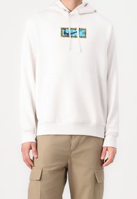 Vit huvtröja med framfickan, förlåtande och med en färgglad grafisk design på en rektangulär lapp. Tillverkad av mjukt tyg.